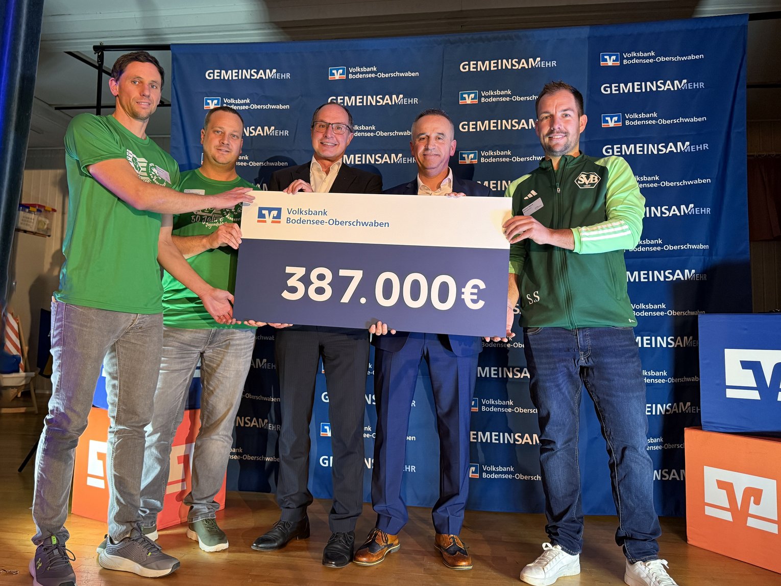 Fünf Männer stehen auf der Bühne und präsentieren einen Scheck über 387.000 Euro
